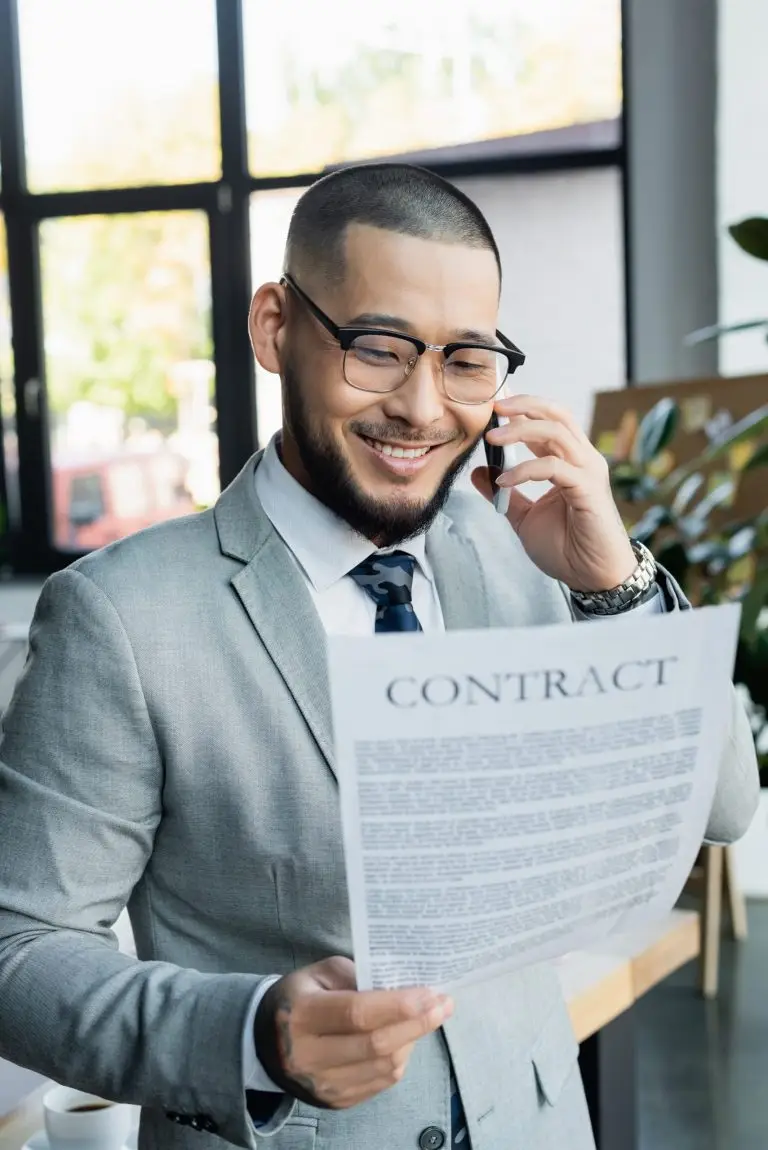 happy asian businessman looking at contract during phone conversation in office